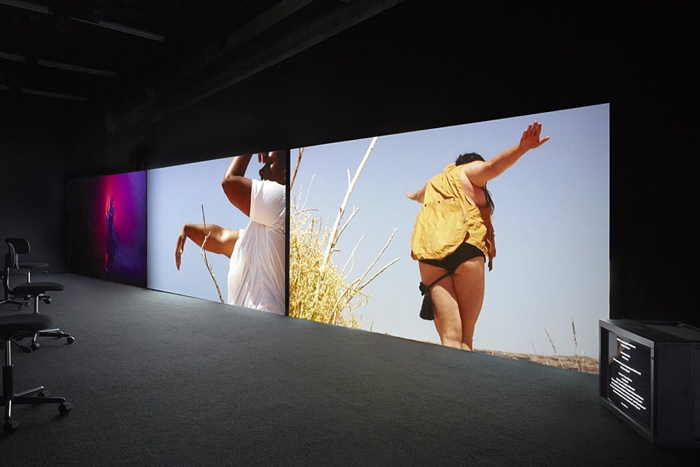 A.K. Burns, installation view of A Smeary Spot, 2015, four-channel video installation, dimensions variable (artwork © A.K. Burns, image provided by the artist, Callicoon Fine Arts, NY, and Michel Rein Gallery, Paris/Brussels).Videos 1–3: HD color, six-channel sound, TRT 53:13 minute synchronized loop; video 4: SD b/w, silent, TRT 4:00 minute loop. Videos 1–3 are each projected onto 81 x 144 in. (205.7 x 365.7 cm) walls, built at an angle to the gallery walls. Gallery is painted black with black industrial carpeted floor and twelve office chairs for seating. Video 4 is presented on a separate 29 in. (73.6 cm) Sony cube-monitor. 