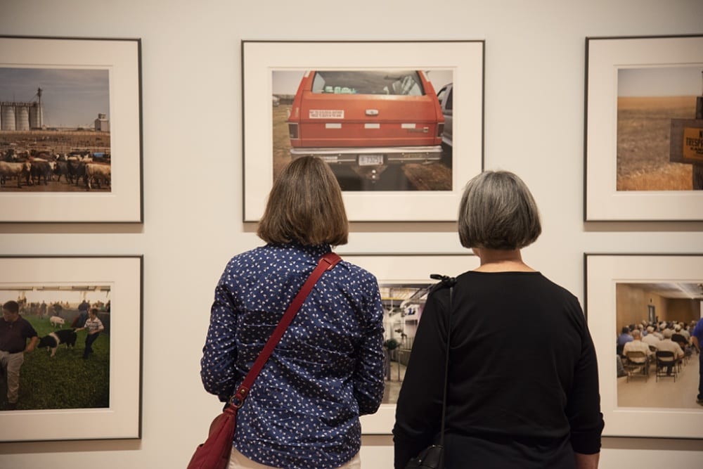 Two women are standing in front of the exhibition "Larry Schwarm: Kansas Farmers." Both women have their backs to the photographer, yet we can see that the woman on the right is wearing a black, long-sleeved sweater, the woman on the left is carrying a red purse. Additionally, the woman carrying the purse is wearing a blue-and-white polka-dot, long-sleeved shirt. Within this frame, there are a total of six images on display, all photographs of Kansas farming, from landscapes to public hearings. Some photographs are populated with people, while others include animals, machinery, and cars.