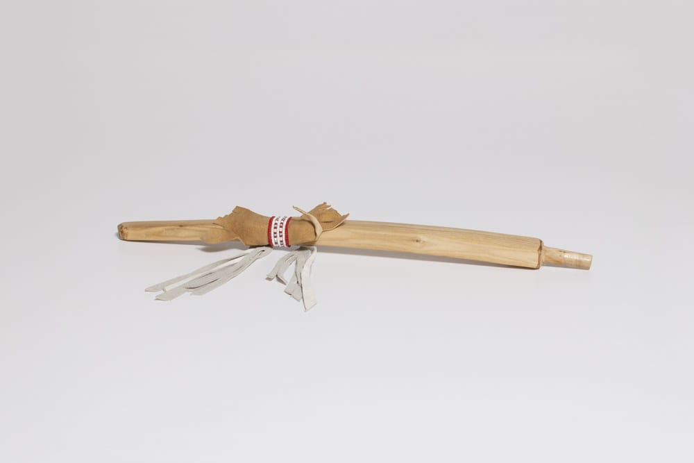 The stem of an Indigenous pipe. The pipe is the color of a light-toned, natural wood with a ribbon and tassels attached (red, white, and green).