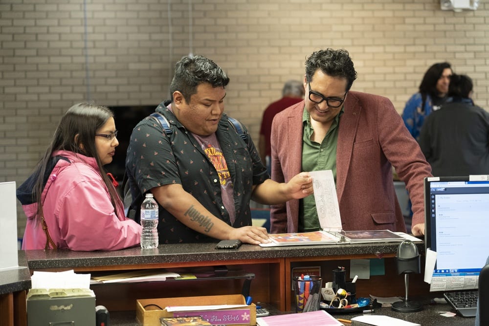 Three people are standing at the circulation desk in the Haskell Indian Nations University library. Adrian Stimson stands on the right in conversation with two students. There is a photo album on the desk, and as Stimson looks through it, the two students next to him, a man and a woman, guide his gaze by pointing out interesting details. The middle figure in the photograph, the young man, lifts a page of the album with his left hand while his right one rests on the circulation desk. All three people look downward at the album as Stimson smiles. On the circulation desk itself we see a computer screen, writing utensils, a plastic water bottle, and a cell phone.