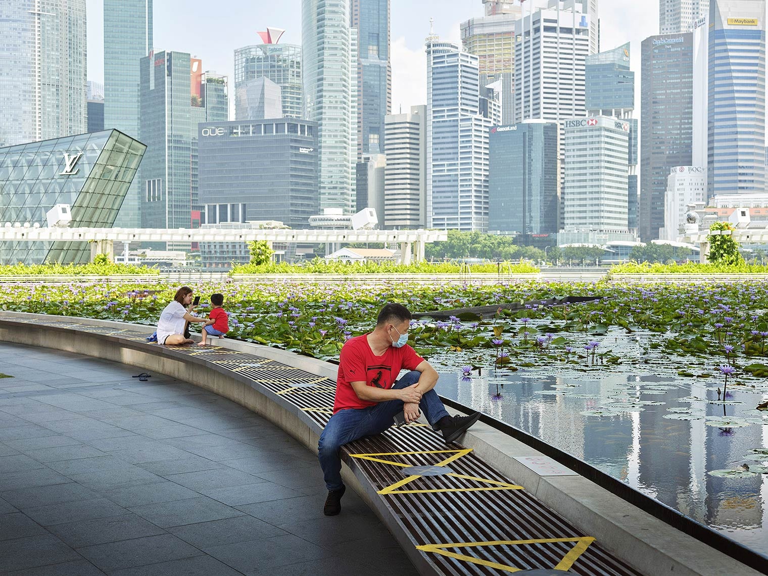 A person wearing a surgical mask sits on a bench by a lily pond in front of Singapore's skyline