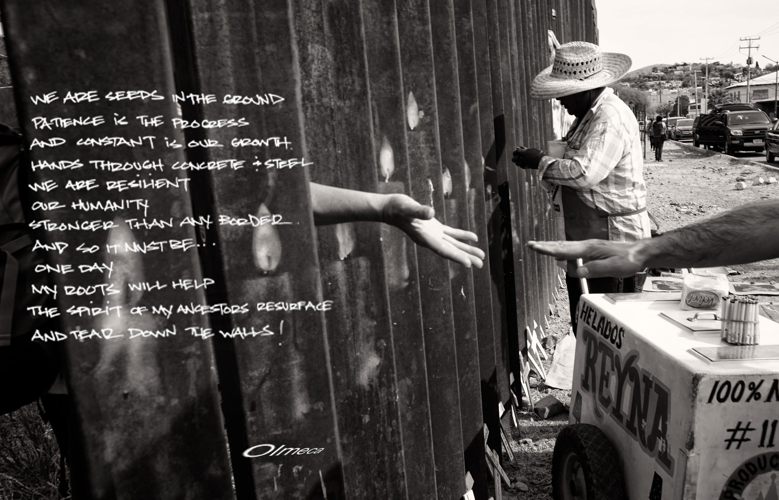 Black and white photo of the US-Mexico border wall built by the United States. A hand strains to reach out through the left-hand side of the image/wall; another hand reaches to meet it from the right-hand side of the border/image. A poem is superimposed over the image's left side.