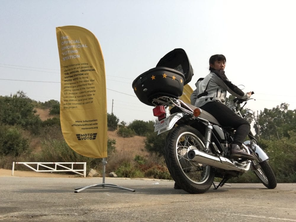 The artist Carol Zou on her motorcycle with a ballot box on the back of the bike, next to a pop-up flag giving info about the voting station