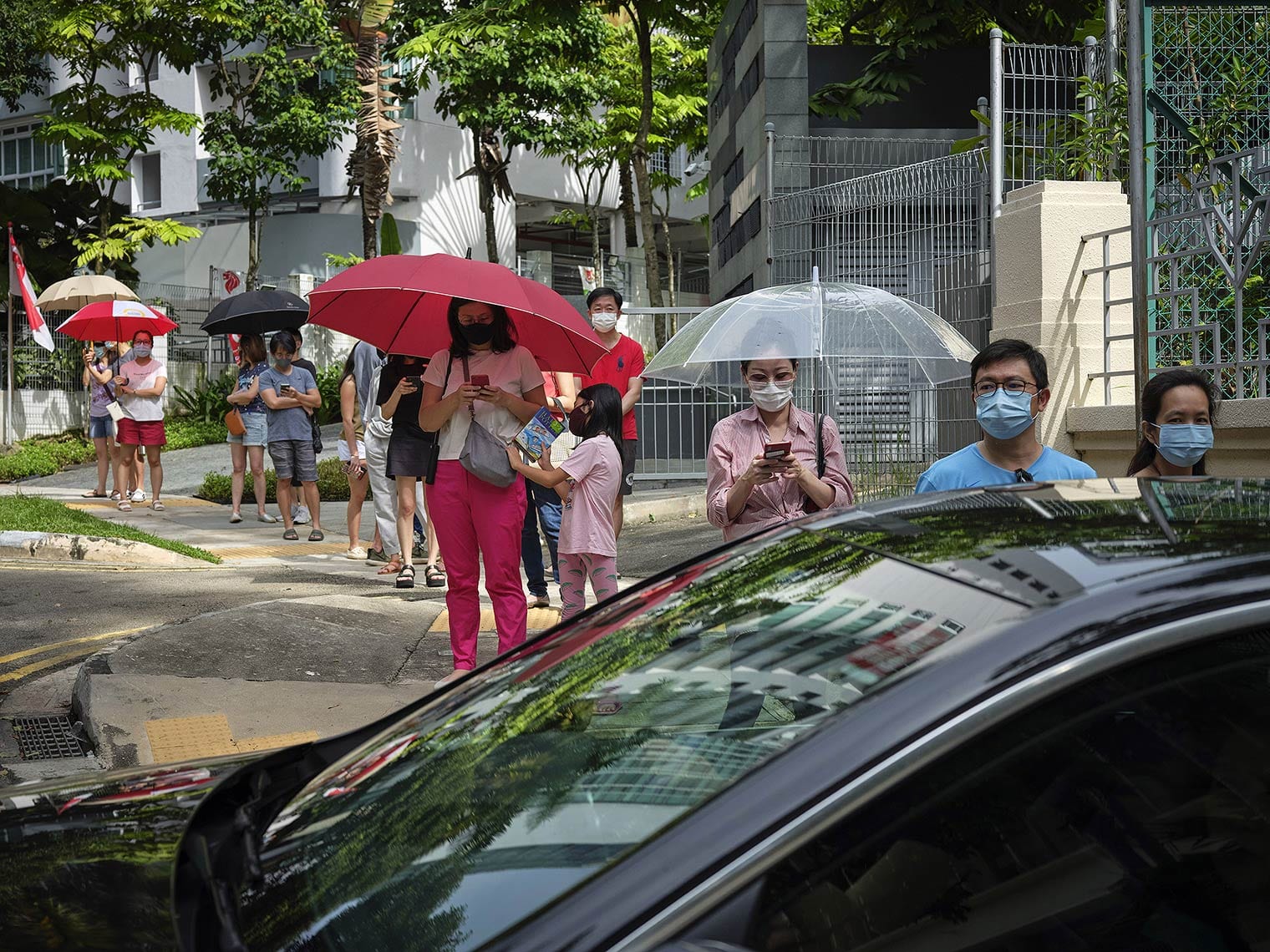 People wearing masks and standing under umbrellas wait in a socially distanced line on a tree-lined city street