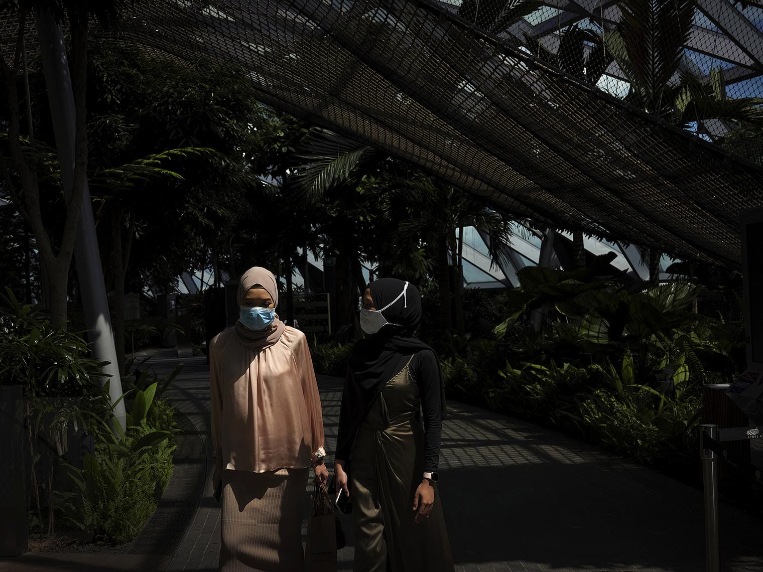 Two masked women walk under a mesh roof, surrounded by greenery