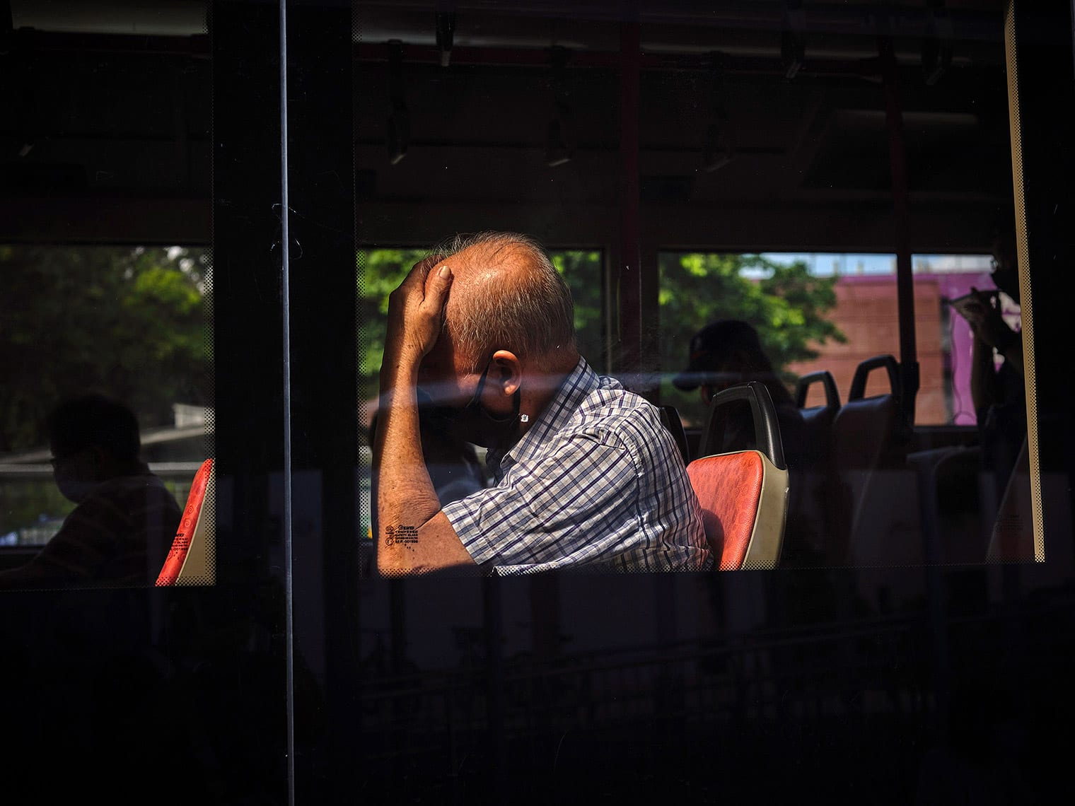 Masked man leans against glass window of a bus, cradling his head