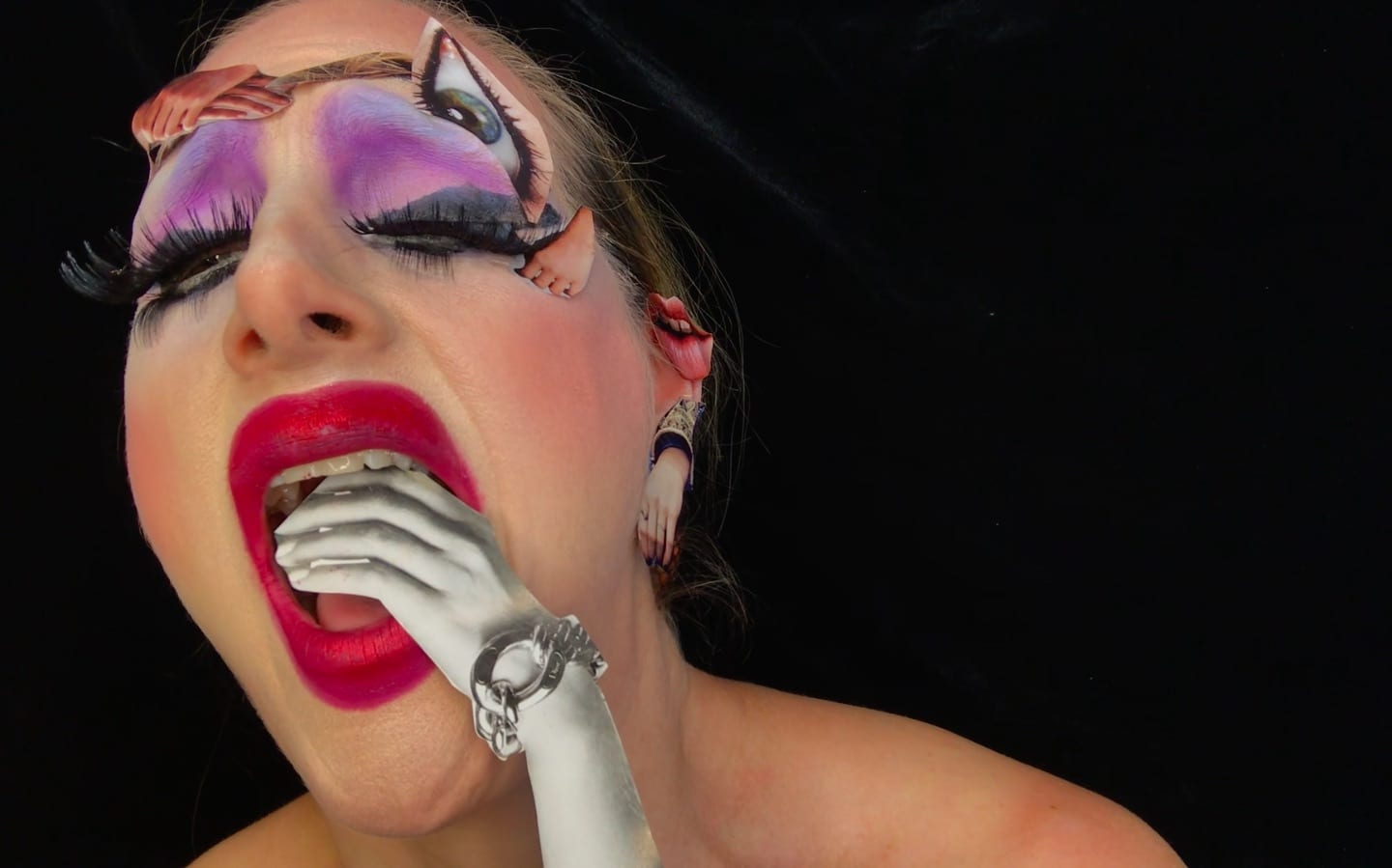 Color video still of performer Monique Jenkinson performing as her drag persona, Fauxnique. She wears heavy makeup, devouring an image of a hand wearing a large bracelet that appears to have been cut from a fashion magazine; magazine cutouts of other body parts are stuck to her forehead.
