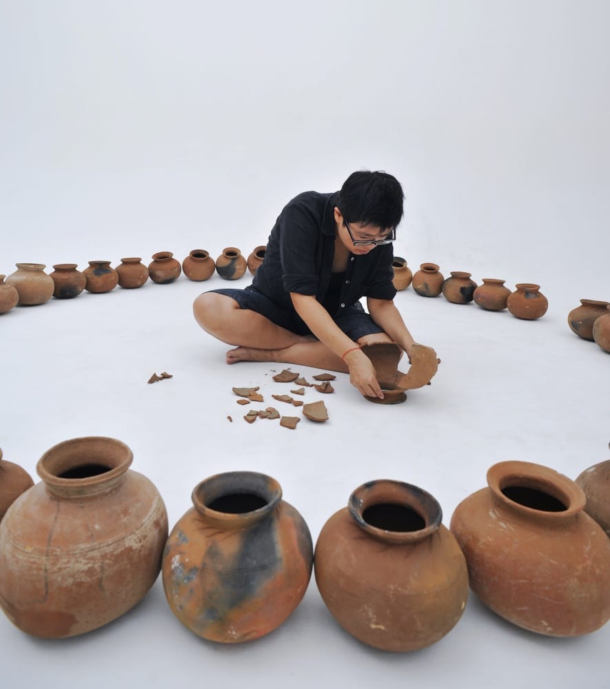 Color photograph of forty clay pots arranged in a circle. A woman dressed in black sits in the middle of the circle. In her left hand, she holds a broken clay pot.