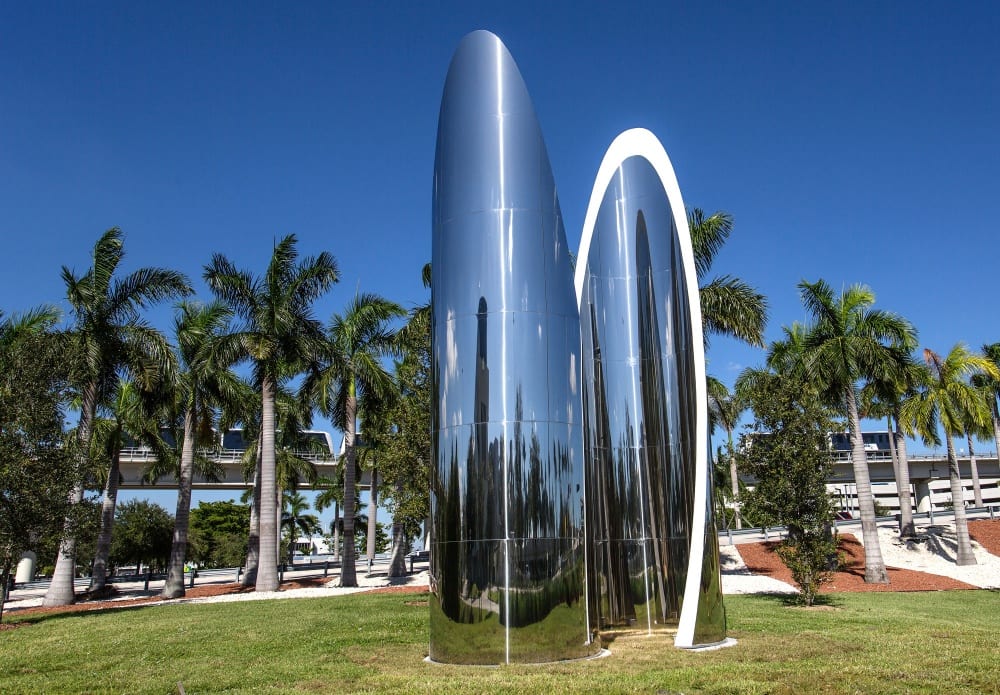 A public sculpture by Fred Eversley, shown on a grassy space against a backdrop of palm trees and blue sky. Two large cylindrical stainless steel forms are sliced on a diagonal (forming parabolas) and overlapped. Neon white light borders the edge of one cylinder.
