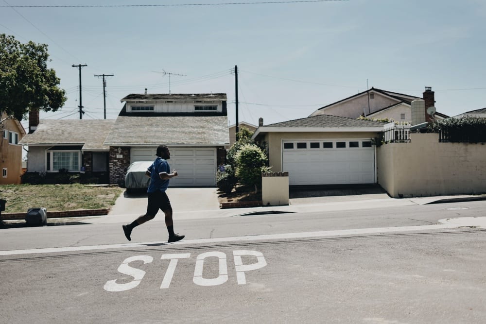 A man runs along a sidewalk in the bright sunlight with houses behind him and the word STOP painted on the ground