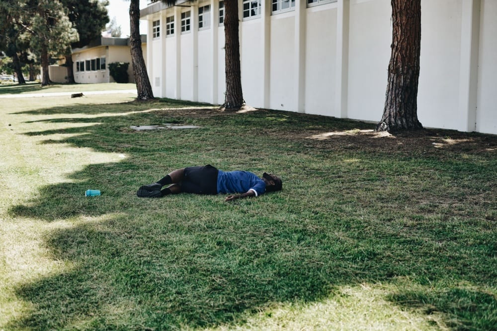 A man lies on a lawn, looking upward, with a large white building and trees behind him