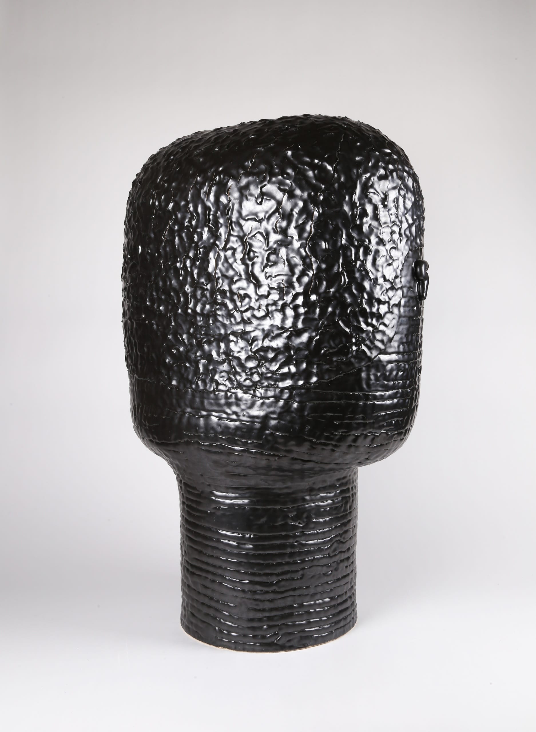 Color photograph with a back view of an abstract ceramic portrait head that appears to be made from a long coil of clay. The head is sculpted to appear square or block-like. The photo shows the rear of the portrait, detailing the texture of the figure's short cropped hair. The object is monochromatic (black), with a slight sheen to the glaze.