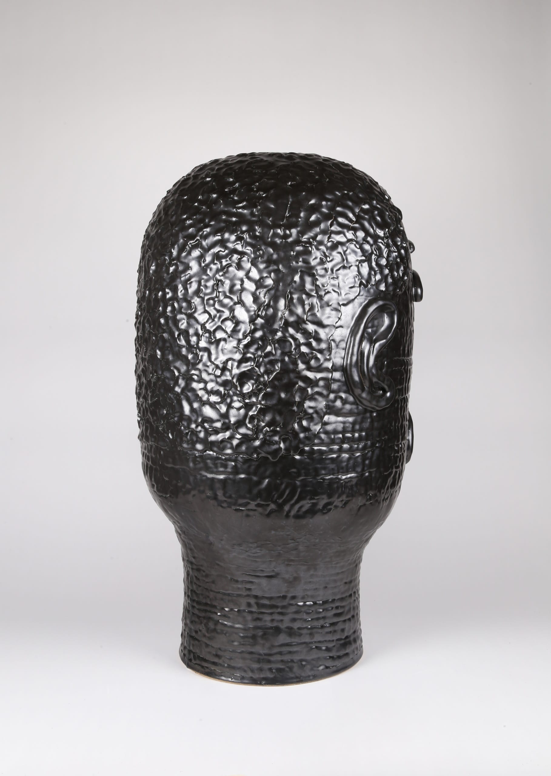 Color photograph with a back view of an abstract ceramic portrait head that appears to be made from a long coil of clay. The rear of the portrait details the texture of the figure's short cropped hair. The object is monochromatic (black), with a slight sheen to the glaze.
