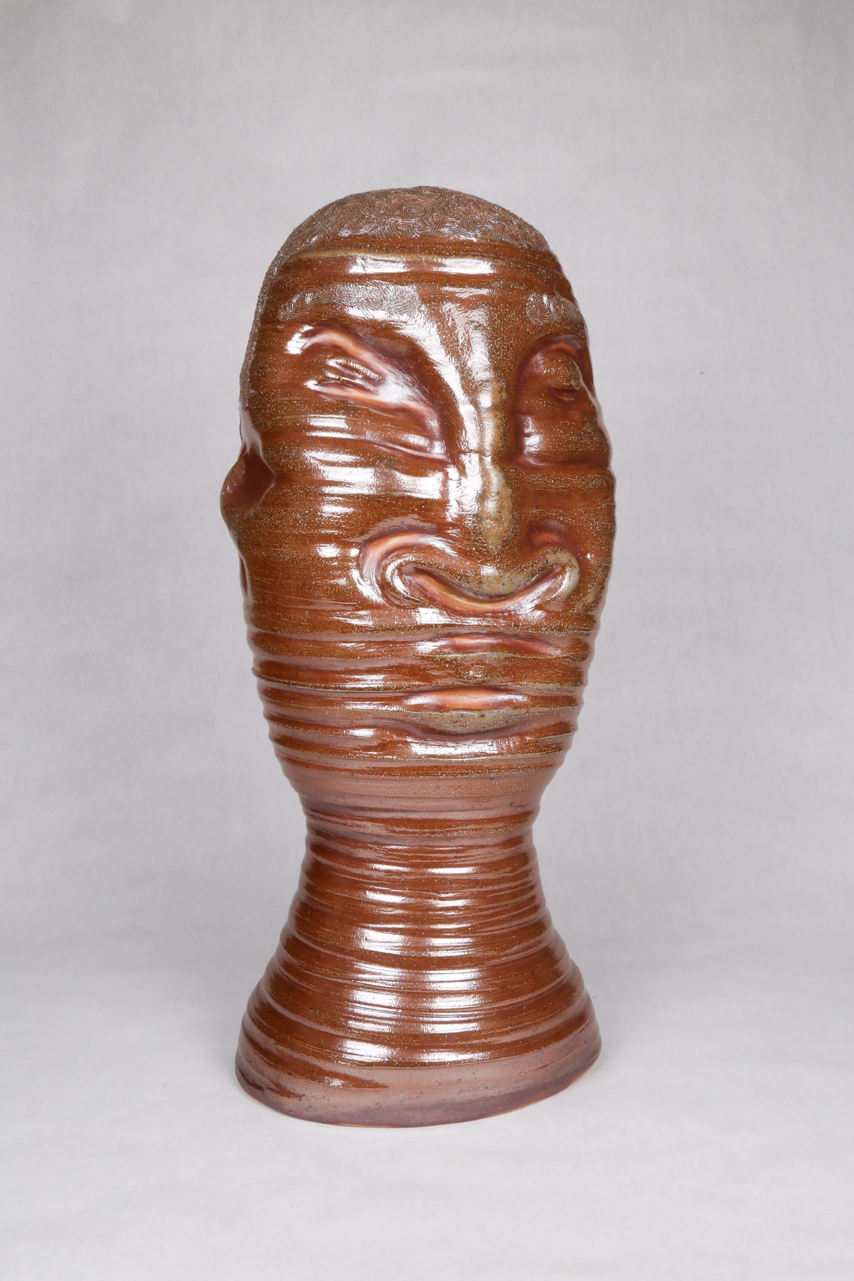 Color photograph with a frontal view of an abstract ceramic portrait head, with distinctive ridges wrapping around the circumfrance of the head. The object is monochromatic (warm brown) with a light, luminescent glaze applied to its surface. It appears as though it was made on a spinning potter‘s wheel.
