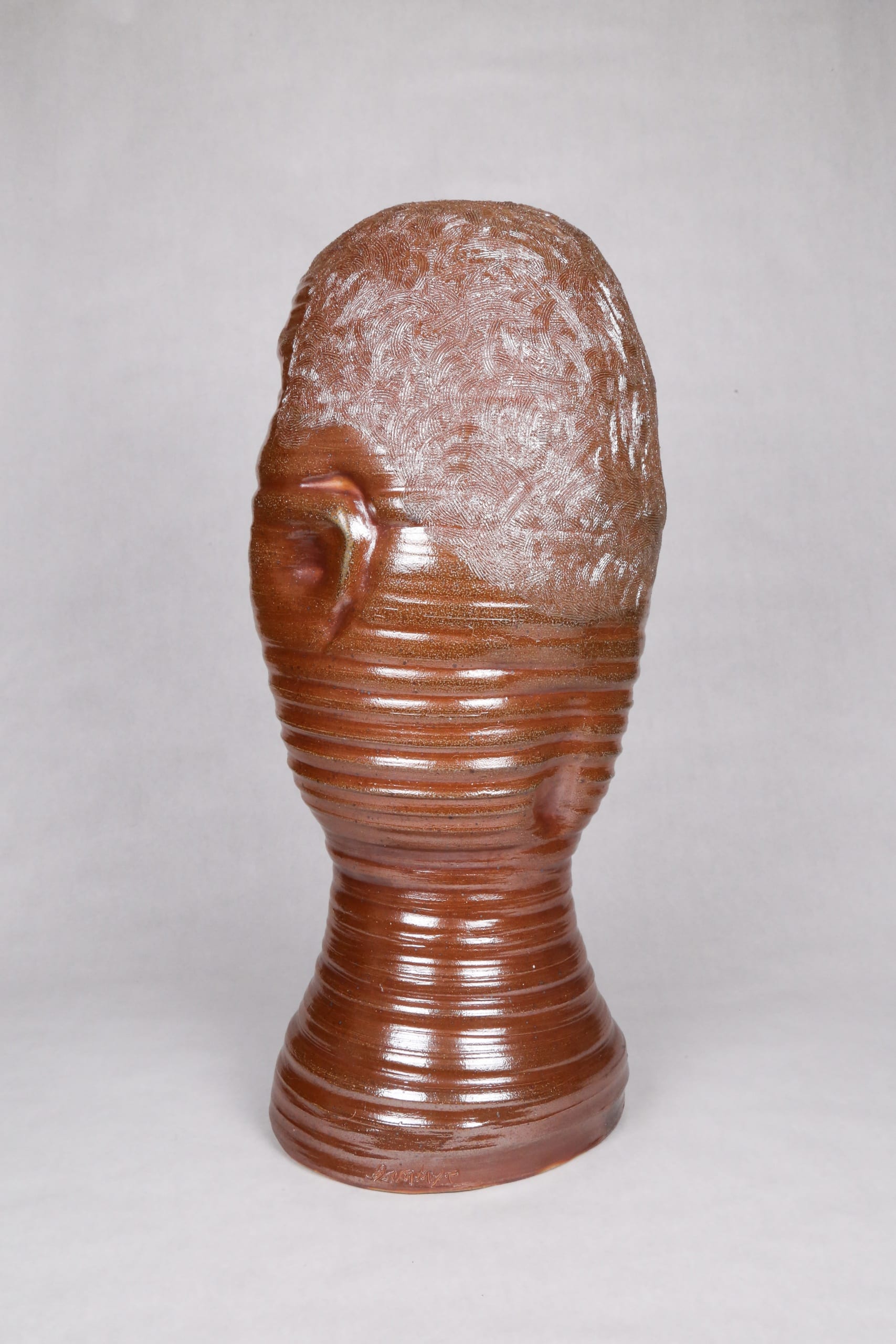 Color photograph with a back view of an abstract ceramic portrait head with distinctive ridges wrapping around the circumfrance of the head. The object is monochromatic (warm brown) with a light, luminescent glaze applied to its surface. It appears as though it was made on a spinning potter‘s wheel.