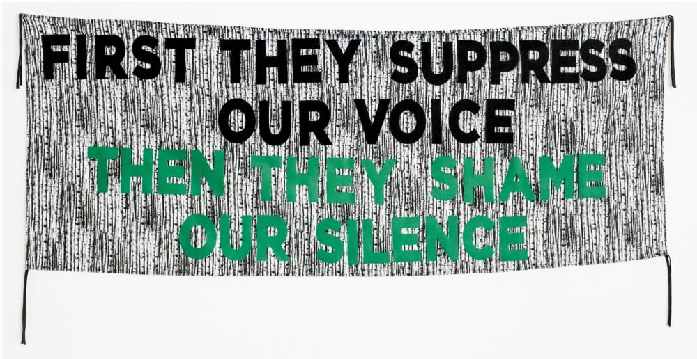 A large horizontal banner with a black-and-white-streaked background reads, in bold black letters, "FIRST THEY SUPPRESS OUR VOICE." Then, in bold green letters directly beneath, "THEN THEY SHAME OUR SILENCE."