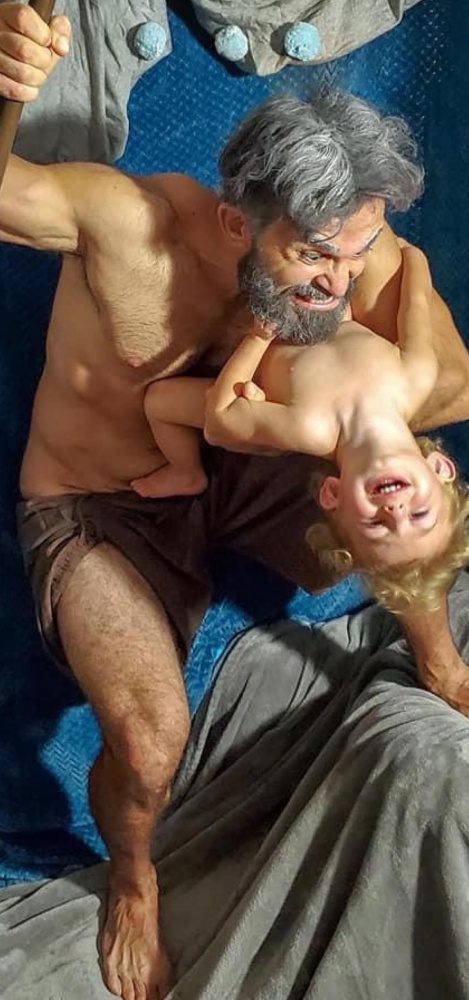 A gray-haired man dressed only in a towel loincloth looks fiercely at a young child draped over his arm. This image is a re-creation of Rubens's Saturn Devouring His Son. In this version, the child appears upside down and laughs with delight. This re-creation is adorable because the young child is having so much fun!