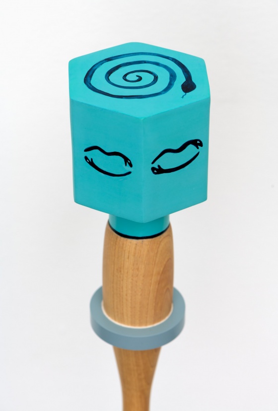 Detailed view of the top of a blue, hexagonal ceramic rattle with a wooden handle, revealing the image of a line drawing of a coiled snake on the top of the rattle.