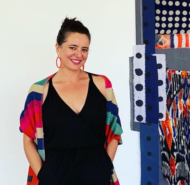Color photograph of Leeza Meksin, smiling and looking toward the camera while standing next to an artwork