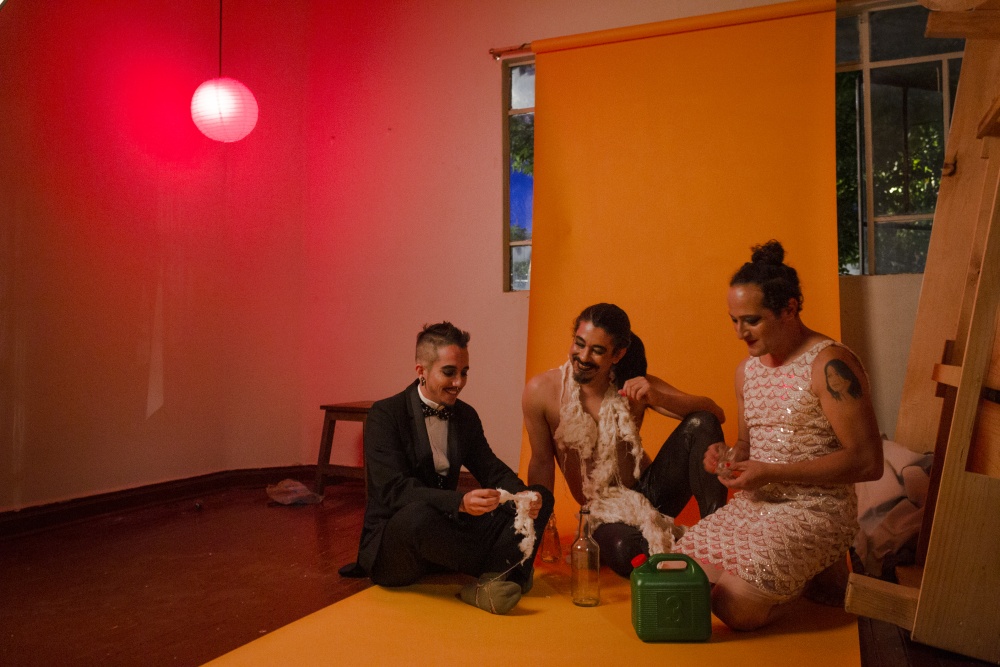 A transgender man, a nonbinary person (the artist), and a gender-nonconforming person are seated on a roll of orange photographer’s paper that hangs from the wall behind them. In front of the individuals are an empty glass bottle and a gas canister. The transgender man pulls apart cotton while the nonbinary person wears unprocessed cotton around their neck.