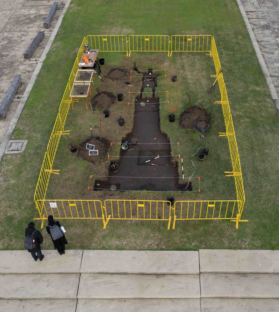 A sculptural trench by the artist Nicholas Galanin, in the shape of a shadow cast by a monument of explorer James Cook, is dug into a grassy plaza and surrounded by yellow fencing.