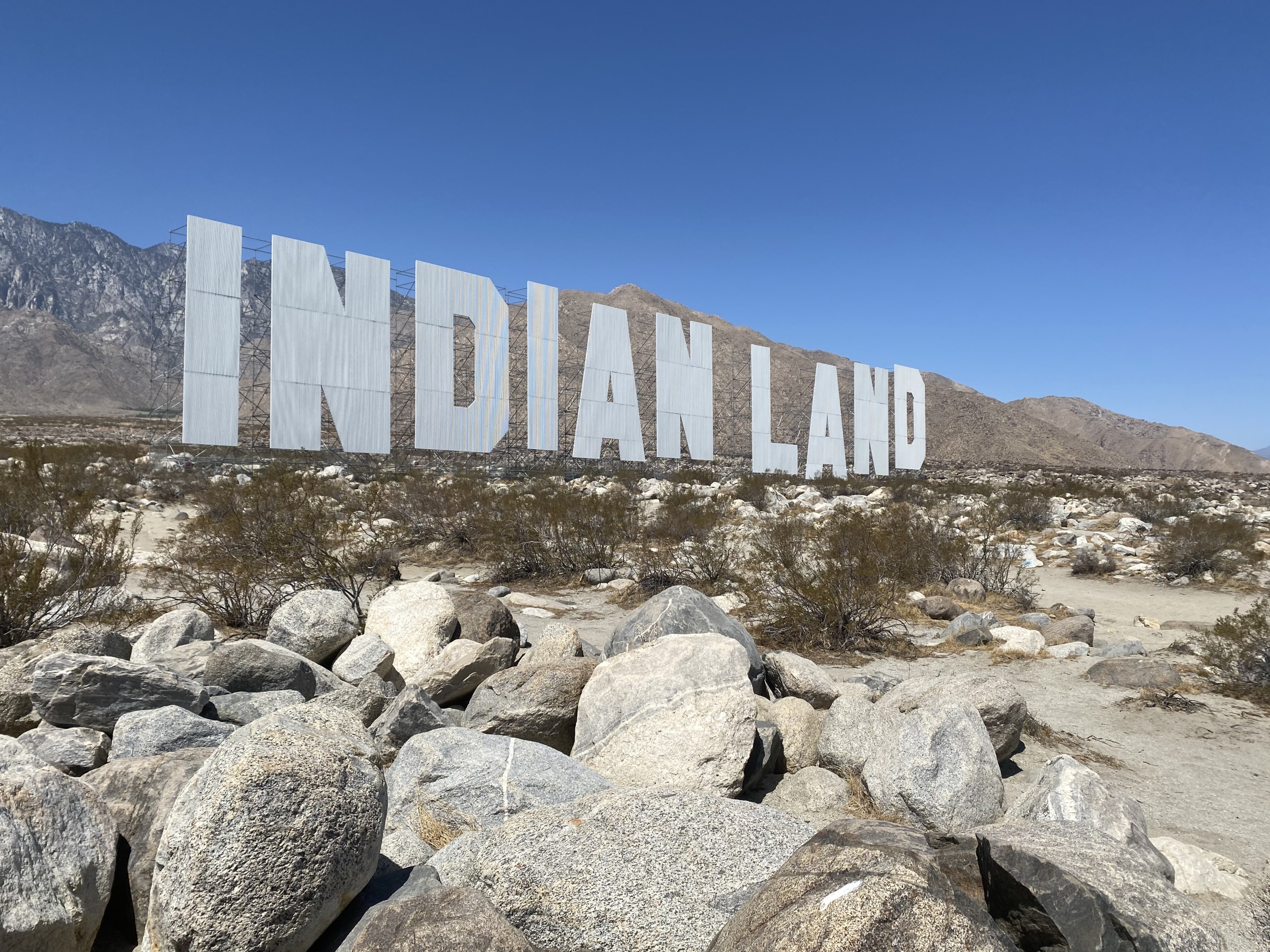 A large-scale installation resembling the Hollywood sign but spelling out “INDIANLAND,” by artist Nicholas Galanin, installed among boulders with desert mountains in the background.