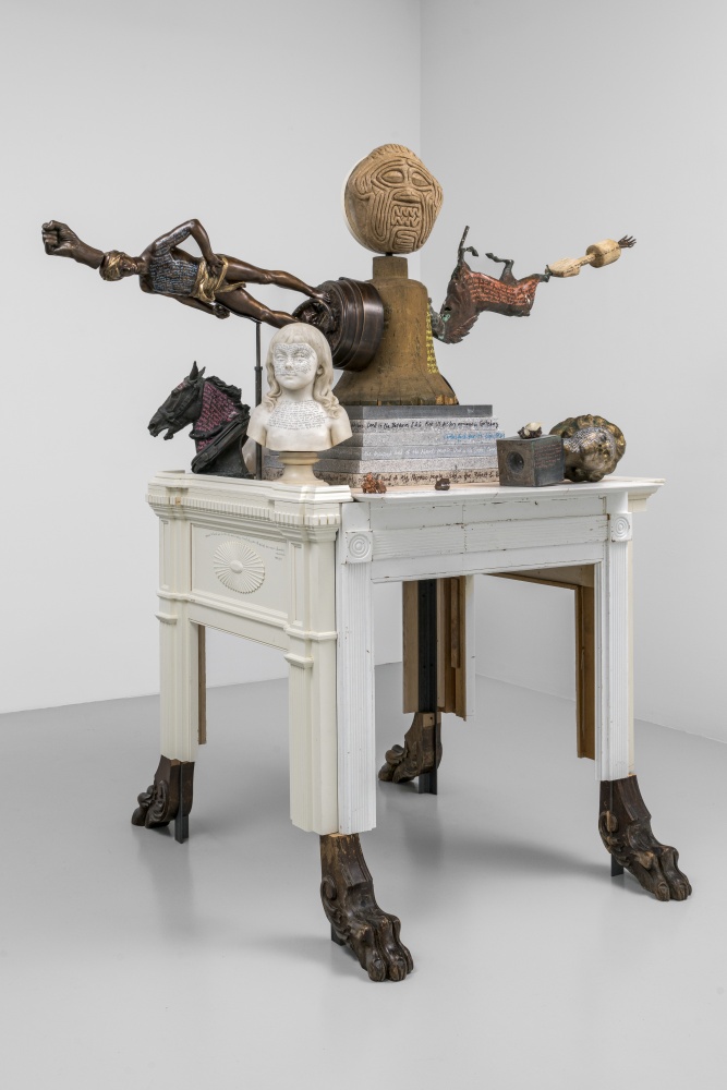 A photograph of a sculpture in a gallery in the shape of an anthropomorphic monster, composed of repurposed objects including a wooden bell form used by a foundry, mantelpieces, wooden lion’s feet from furniture, and small marble and bronze sculptures on which the artist has written.