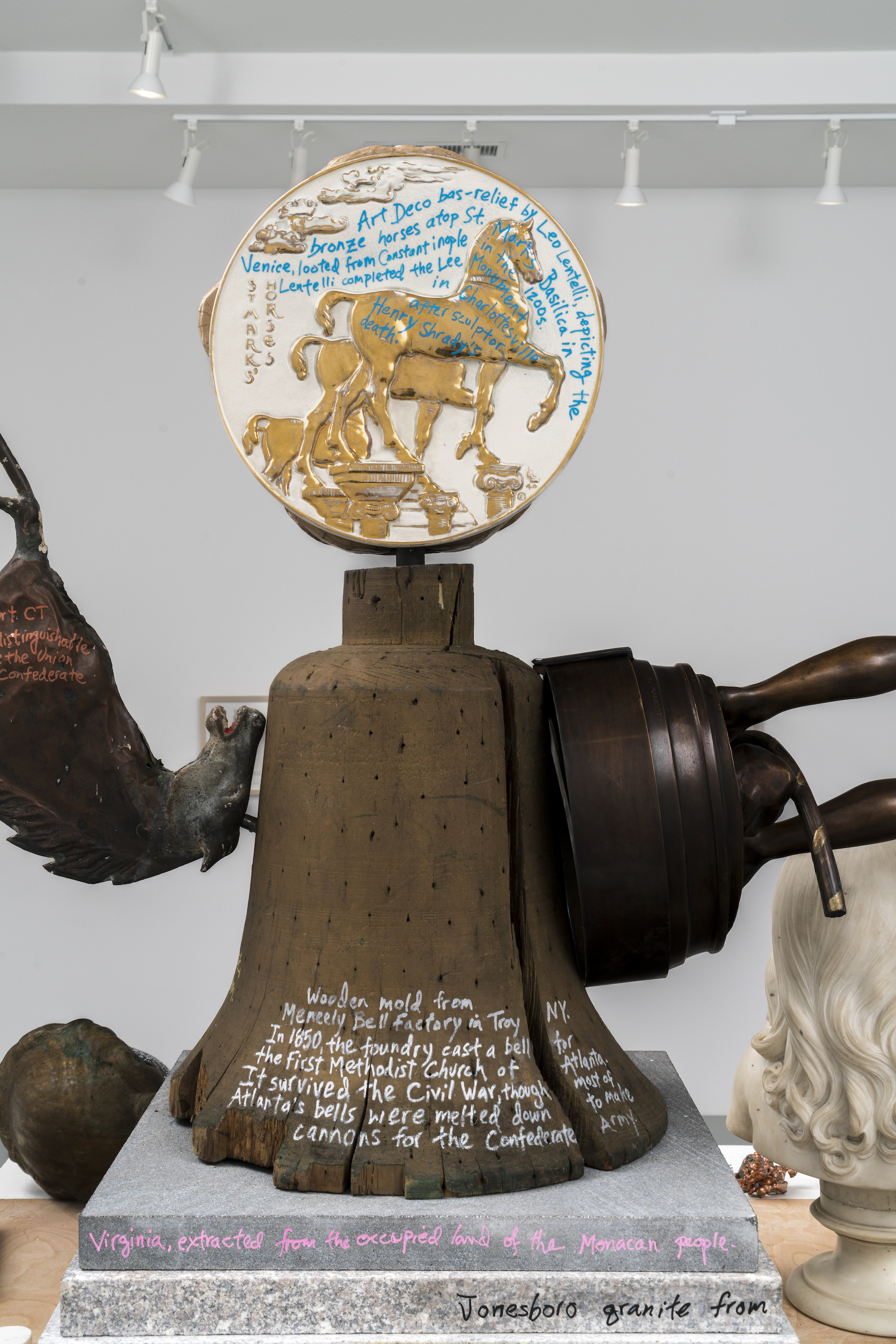 A photograph of a detail of a sculpture in a gallery in the shape of an anthropomorphic monster. The photograph shows the figure’s torso, which is composed of repurposed objects including a wooden bell form and a circular bas-relief. On the bell form, the artist has written “wooden mold from Meneely Bell Factory in Troy, NY. In 1850, the foundry cast a fell for the First Methodist Church of Atlanta. It survived the Civil War, though most of Atlanta’s bells were melted down to make cannons for the Confederate Army.” On the circular bas-relief, the artist has written “Art Deco bas-relief by Leo Lentilli, depicting the bronze horses atop St. Mark’s Basilica in Venice, looted from Constantinople in the 1200s. Lentelli completed the Lee Moniument in Charlottesville after sculptor Henry Shrady’s death.”