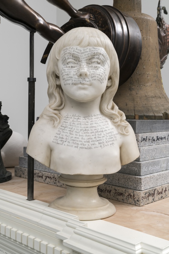 A photograph of a detail of a sculpture composed of repurposed objects. The photograph shows one of these objects, a marble bust of a young girl, on whose face and chest the artist has written “‘Portrait of a Young Girl’ by Thomas Ball, who also sculpted the Emancipation Memorial. Known as the Freedman’s Memorial, it was paid for by formerly enslaved people and depicts Lincoln ‘freeing’ a kneeling, enslaved African American person while holding the Emancipation Proclamation. Ball initially hired a Black model but fired him because of ‘the unpleasantness of being obliged to conduct him through our apartment.’ Ball then used himself as a model but was eventually persuaded by the commission to work from a photograph of a formerly enslaved Black man named Archer Alexander. In a dedication speech for the memorial, Frederick Douglass said: ‘Truth compels me to admit… Abraham Lincoln was not, in the fullest sense of the word, either our man or our model. In his interests, in his associations, in his habits of thought, and in his prejudices, he was a white man.’ Douglass noted that Lincoln was motivated more to save the Union than to free slaves and that he ‘strangely told us that we were the cause of the war.’ In December 2020 the Boston Art Commission voted unanimously to remove the city’s copy of the statue after a public debate determined it reinforced a racist and paternalistic view of Black people.”
