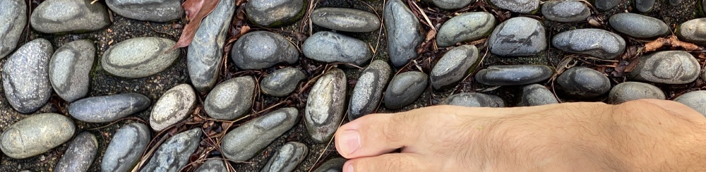 Partial view of the side of a man’s foot standing or walking on ground made up of small, smooth stones, taken from above.