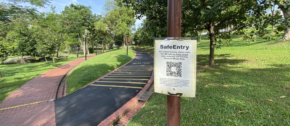 Two intersecting trails, one brick and one asphalt with yellow lines, in a park with bright green grass and many trees. A small “SafeEntry” sign on a brown pole in the foreground includes lots of smaller text and a QR code.