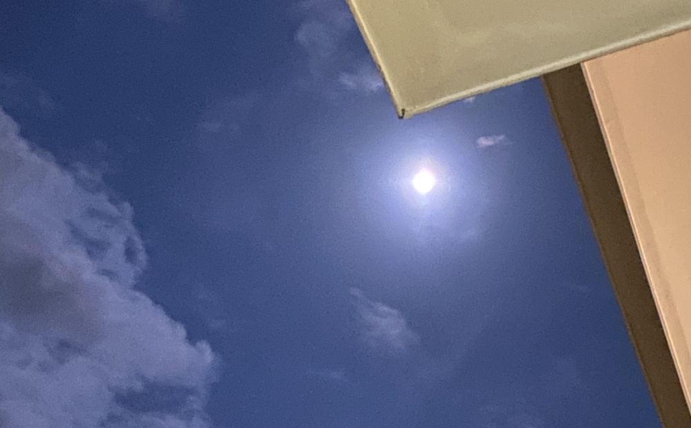 Upward view of blue sky with a small bright sun, clouds, and a part of a building.