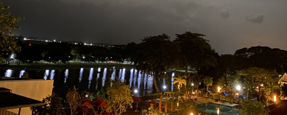 Gray night sky and a body of sea water reflecting the lights illuminating paths in a park with trees on one side and a swimming pool, palm trees, and low buildings on the other.