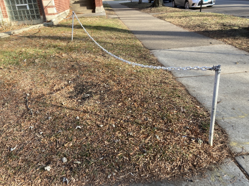 Two metal poles in a yard sparsely covered with brown grass connected by a chain, next to a concrete sidewalk.