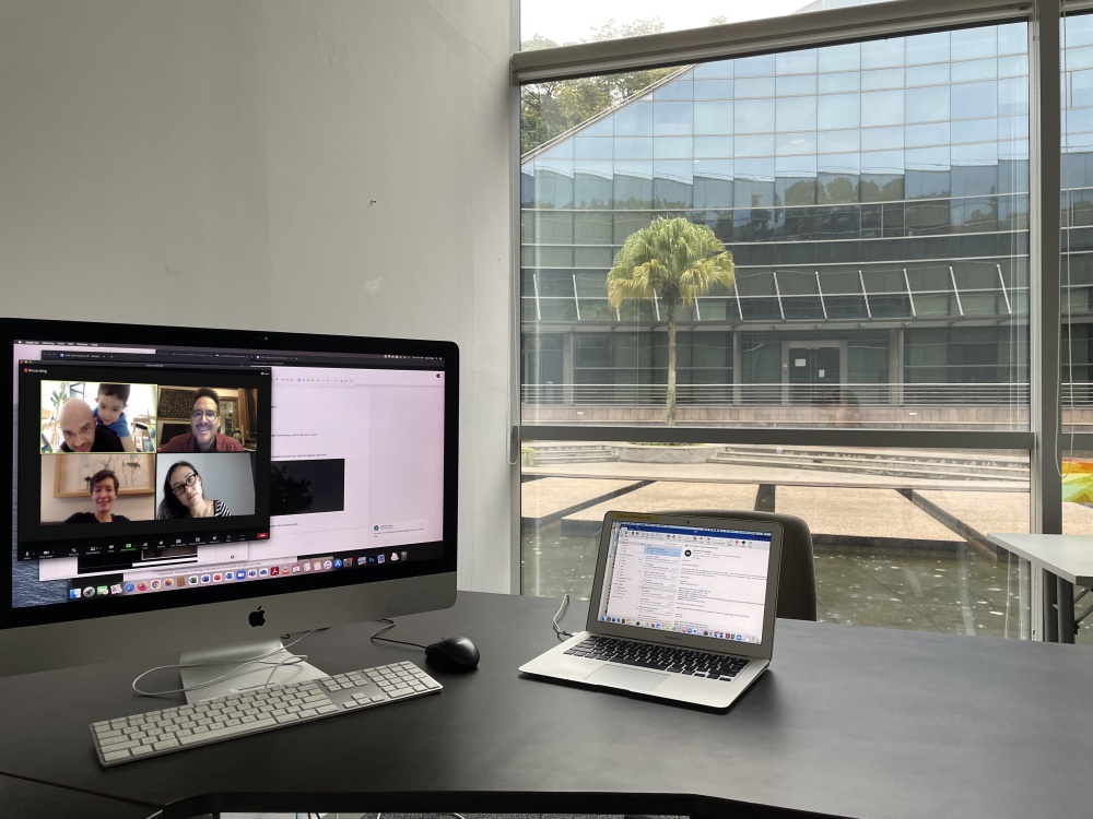 Two computers on a bare desk looking out a large window into a courtyard with square walkways in a reflecting pool. On the left is a large display showing the images of three participants in a video conference, on the right is a smaller laptop computer with documents in progress.