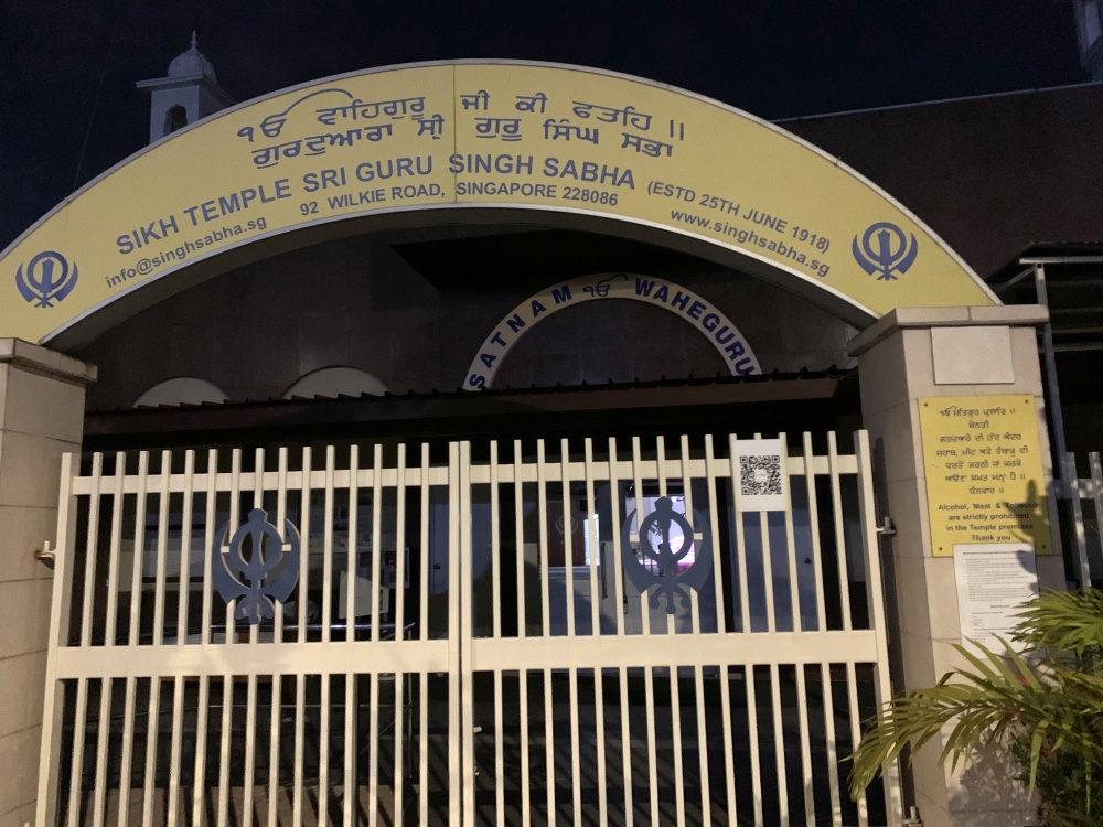 Night view of a closed light-colored gate with a yellow arch with blue symbols and text: “Sikh Temple Sri Guru Singh Sabha.”