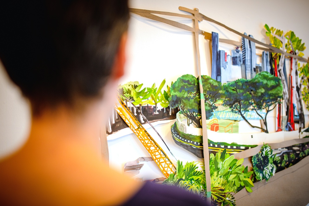 Foreground: out-of-focus image of the back of a person’s head. Middle and background: an installation on or near a white wall including a collaged image made from painted imagery of roads, construction crane, greenery, trees, and skyscrapers.