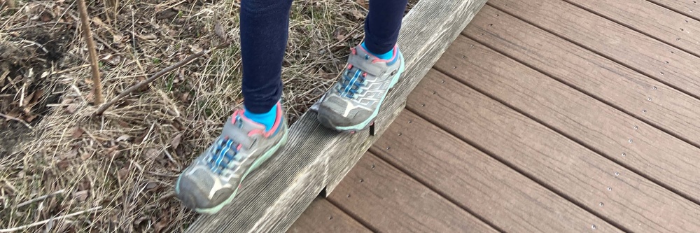 A child’s feet, balancing while walking on a raised concrete curb between a walkway and brown grass, wearing sneakers, turquoise socks, and dark blue leggings. These feet look like they belong to the child in figure 3.