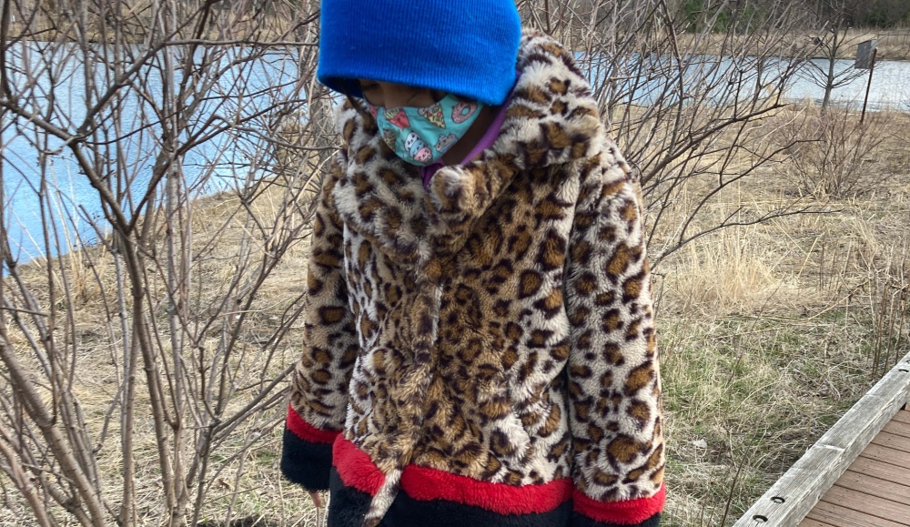 Child wearing a leopard-print coat, turquoise face mask, and blue hat looking down while walking next to brown grass and shrubbery with no leaves.