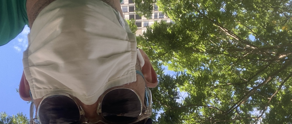 Upward view of the bottom half of a man’s face with a tree with green leaves, blue sky, and a building behind him. He is wearing a white face mask and sunglasses.