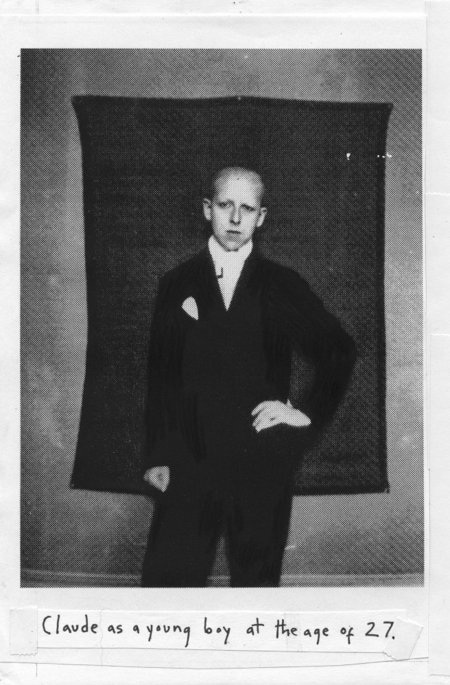 A photograph of a white genderqueer subject with a shaved head, suit, and hand on hip. Hand-written text below it reads in all-caps: CLAUDE AS A YOUNG BOY AT THE AGE OF 27.