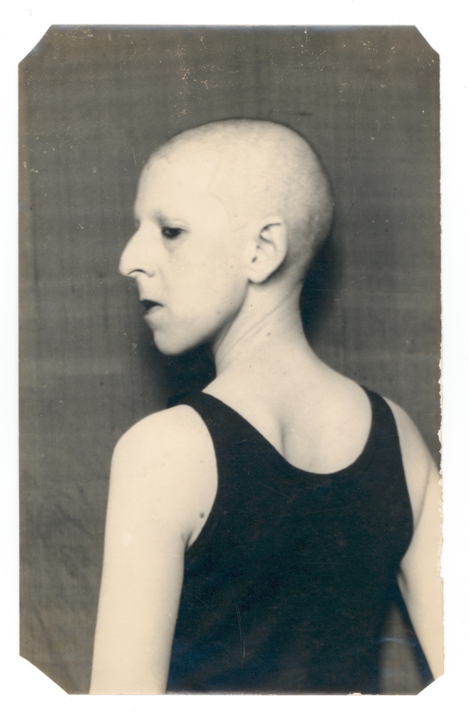 A black-and-white photograph showing a thin white genderqueer Claude Cahun with their back facing the camera and their face in profile. Their head is shaved and they wear a black tank top and stand against a dark background.
