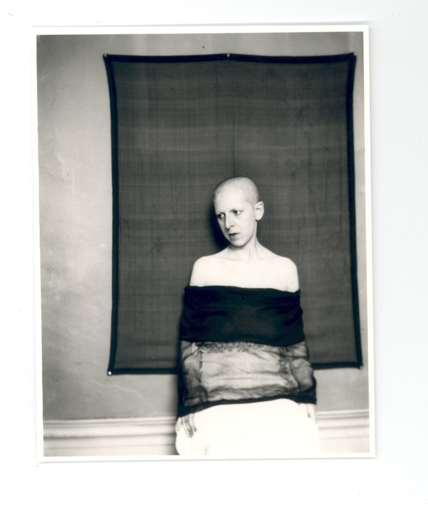 A thin white genderqueer Claude Cahun facing the camera and their face turned to three-quarters profile. Their head is shaved and their shoulders are bare.