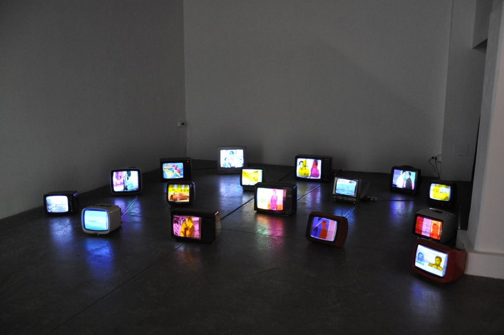 Multiple vintage TV monitors with brightly colored screens are strewn across the floor of a darkly lit white gallery space.