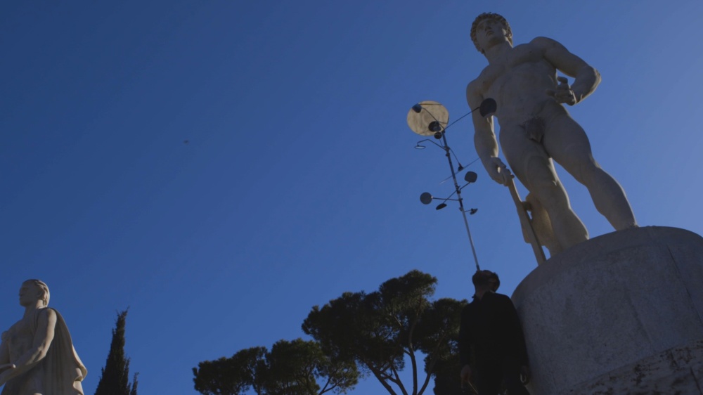 A person, artist Hiwa K, balances on his head a metal pole with attached motorcycle mirrors, near large stone statues of athletes, all seen against a clear blue sky.