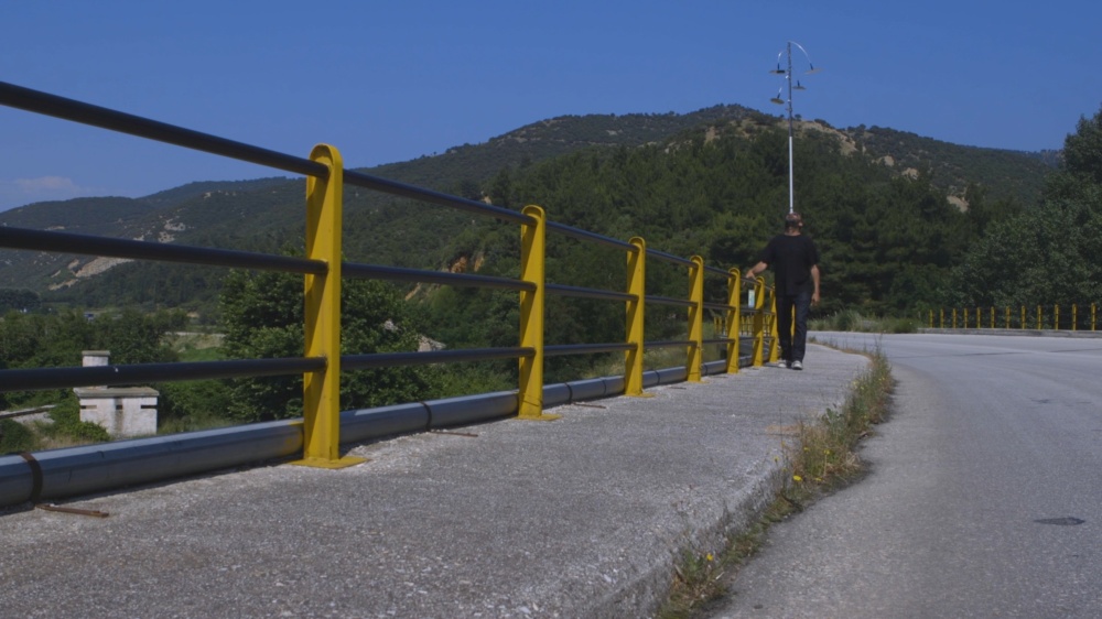 A person walking on the side of a highway, with a natural landscape seen in the background, while balancing on his head a metal pole with attached motorcycle mirrors.