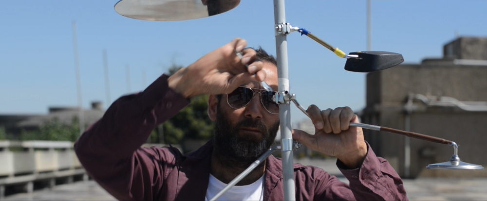 A person, artist Hiwa K, is seen against an urban background, tying motorcycle mirrors to a metal pole.