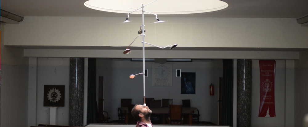 A person balances on his head a metal pole with attached motorcycle mirrors, within a building’s large interior, illuminated from above through a skylight.