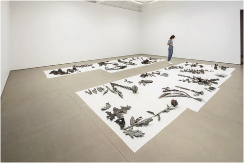 Groupings of bronze sculptures, featuring fragments of plants and flowers, strewn across white sheets of fabric placed directly on a gallery’s floor.