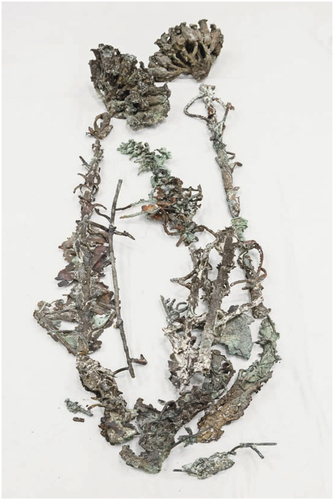 Fragments of an oxidized bronze sculpture, some with green patina, assembled into the shape of a flower plant, resting against a white piece of fabric.